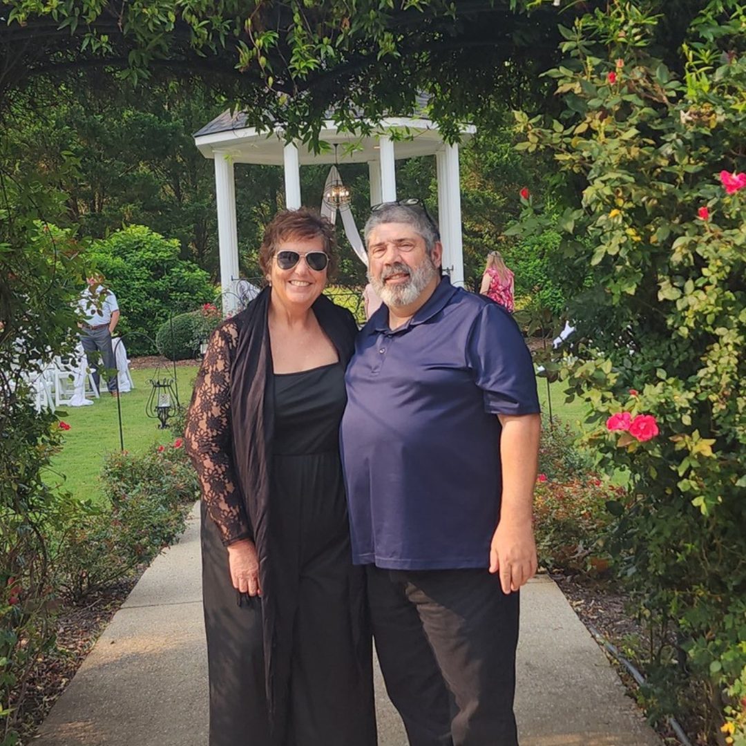 Two people standing on a garden path under a white arbor, surrounded by flowers and greenery.