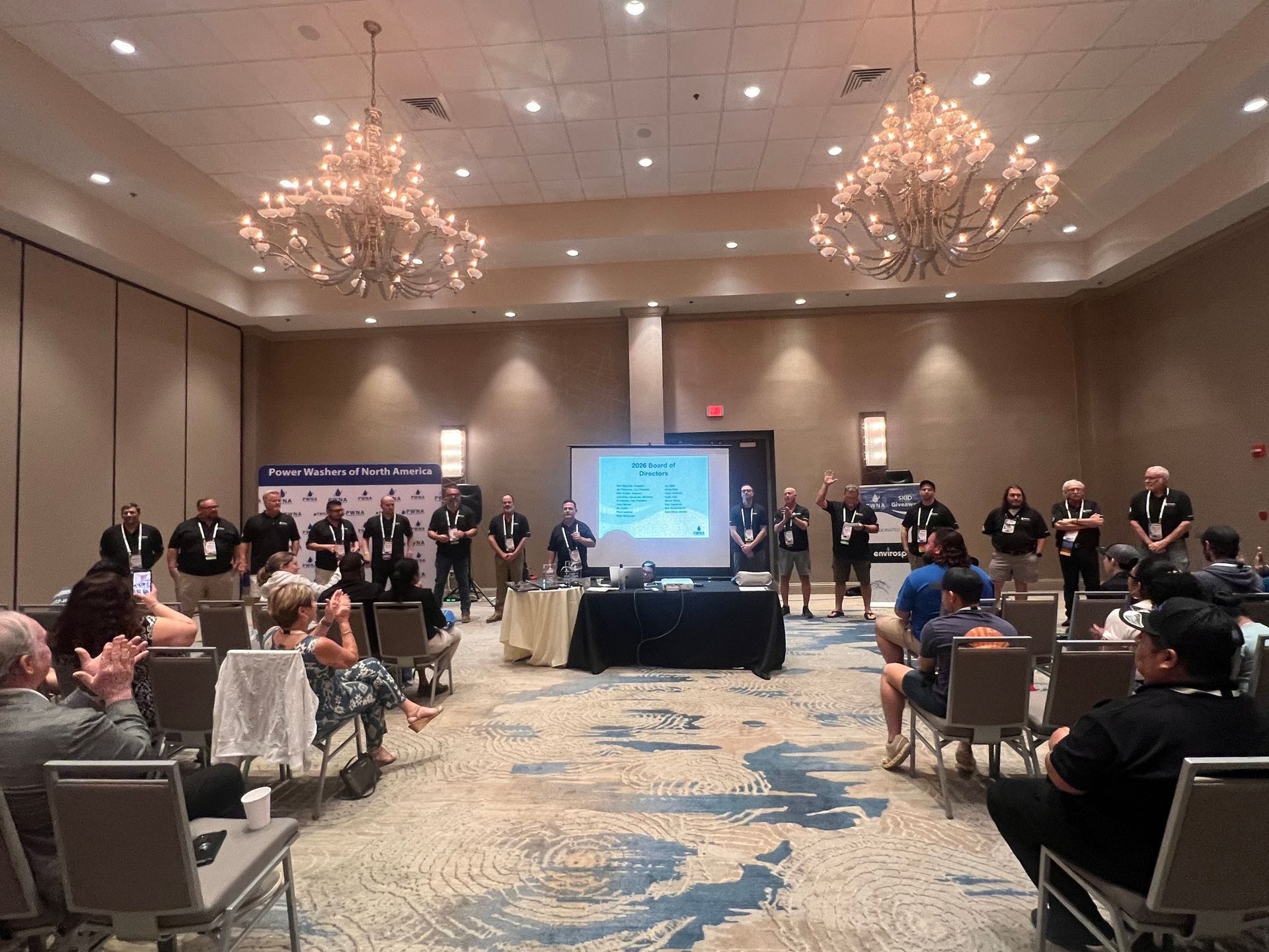 Conference room panel with seated audience, presenters at the front, and a projected screen between two chandeliers.