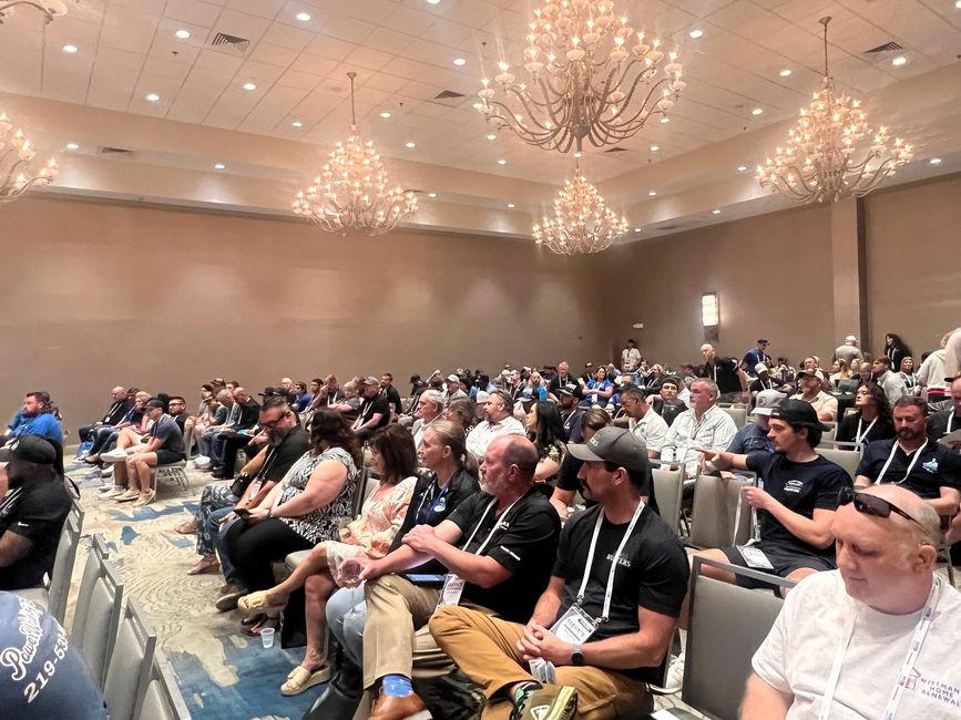 Audience seated in a large conference ballroom with chandeliers and rows of tables and chairs