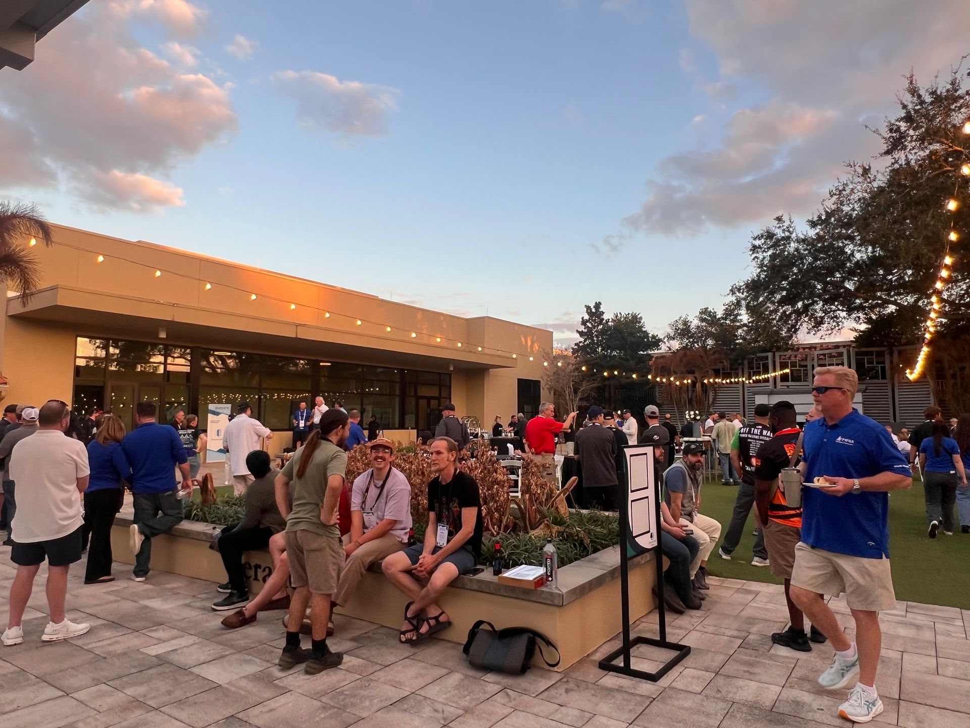 People gathered outdoors at dusk near a building, with a crowd seated and standing on a plaza.