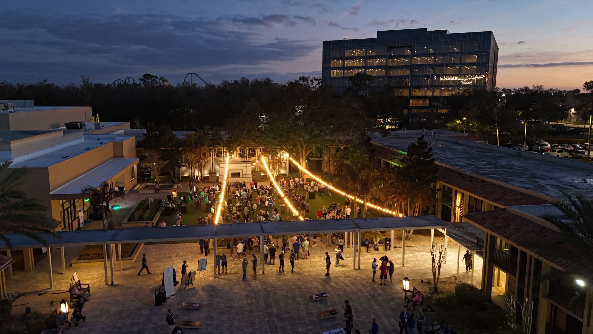 Dusk view of a lit campus plaza with people walking, trees, and a modern building in the background
