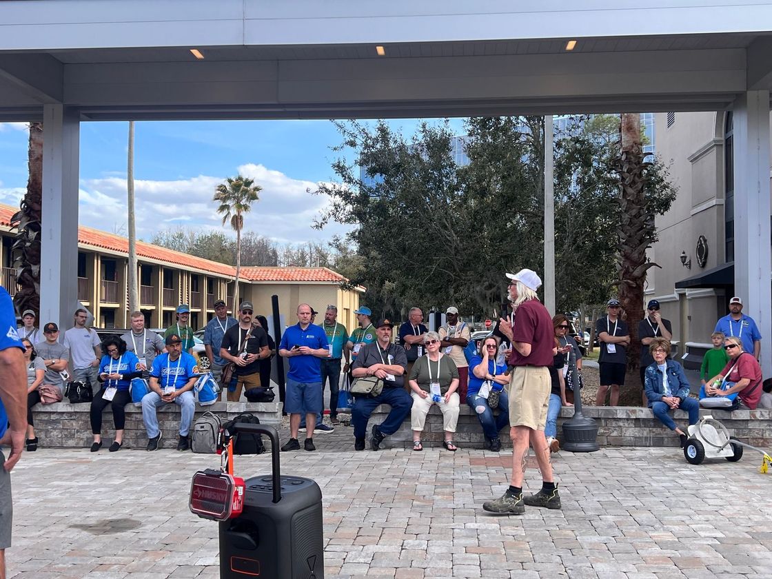 Crowd seated and standing in a covered courtyard, with luggage and a man in maroon shirt in the foreground