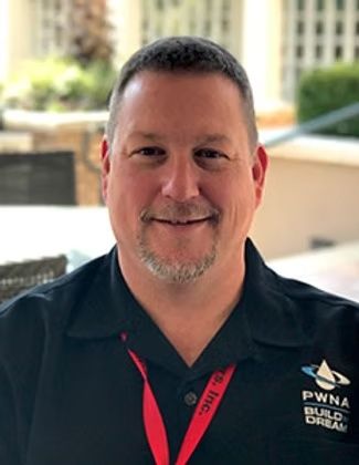 Smiling man in a black shirt with a red lanyard, standing outdoors near a building.