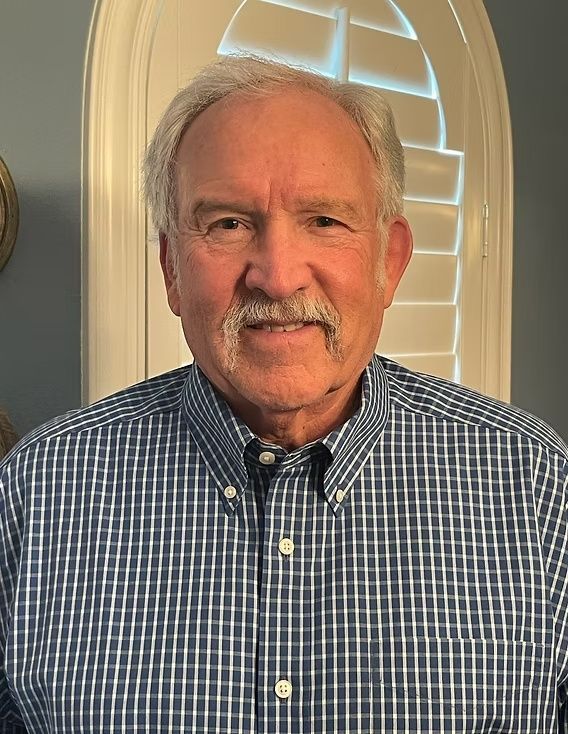Smiling man in a blue plaid shirt standing indoors in front of a window with white blinds