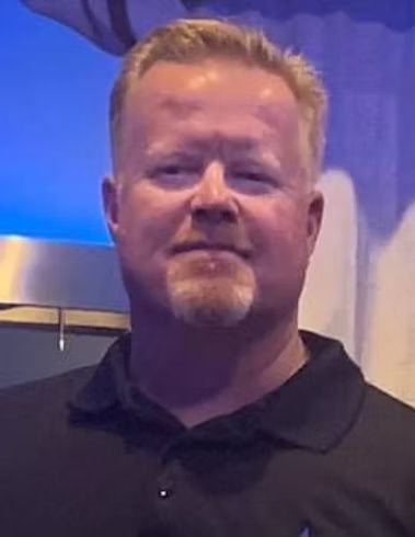 Smiling man in a black polo shirt against a blue-lit indoor background