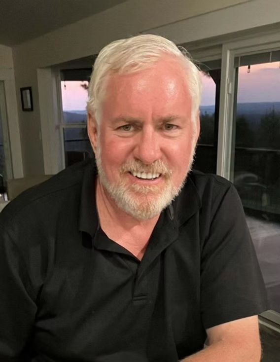 Smiling man in a black polo shirt indoors, seated by a window at dusk.