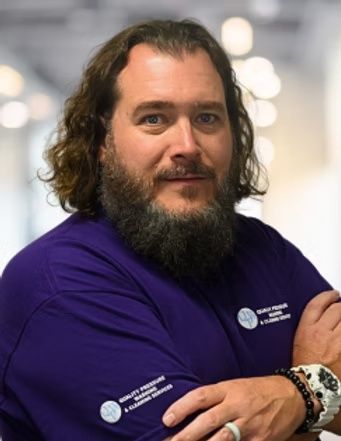 Person in a purple shirt with folded arms, wearing bracelets, against a blurred indoor background.