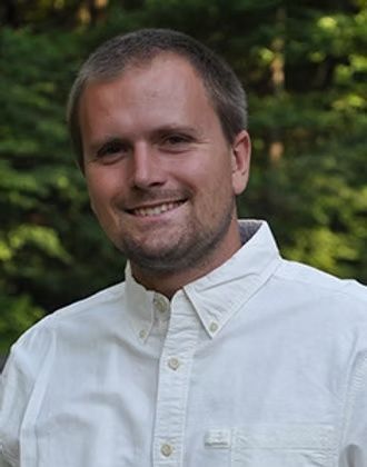 Smiling man in a white button-down shirt standing outdoors with blurred green trees behind him