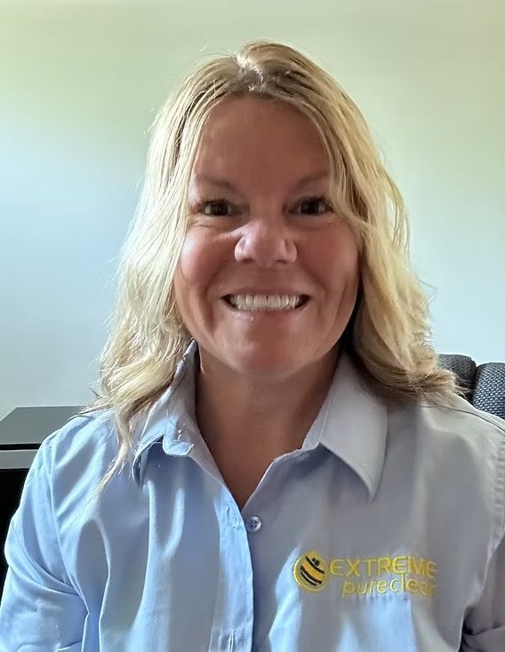 Smiling person in a light blue collared shirt with logo, seated indoors against a light wall background