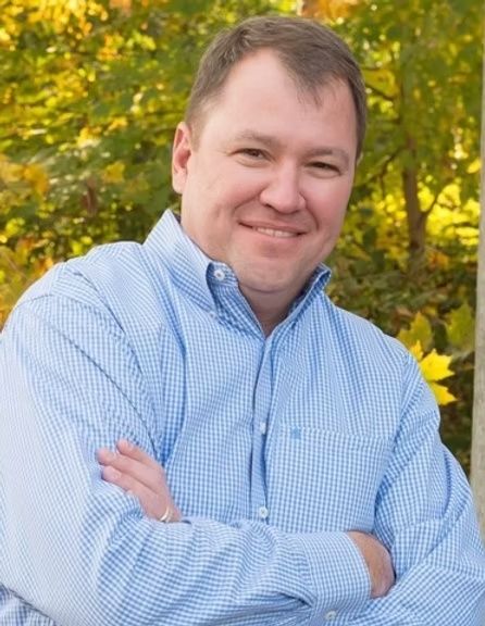 Smiling man in a blue checkered shirt with arms crossed, standing outdoors in front of yellow autumn foliage