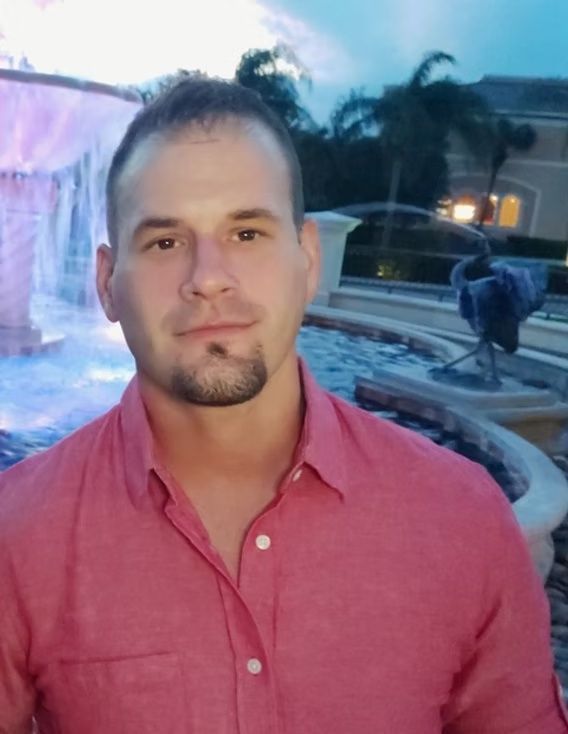 Man in a pink shirt standing outdoors near a lit fountain at dusk.
