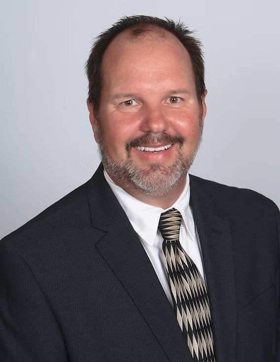 Smiling man in a dark suit and patterned tie against a light gray studio background
