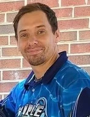 Man in a blue collared shirt smiling in front of a brick wall