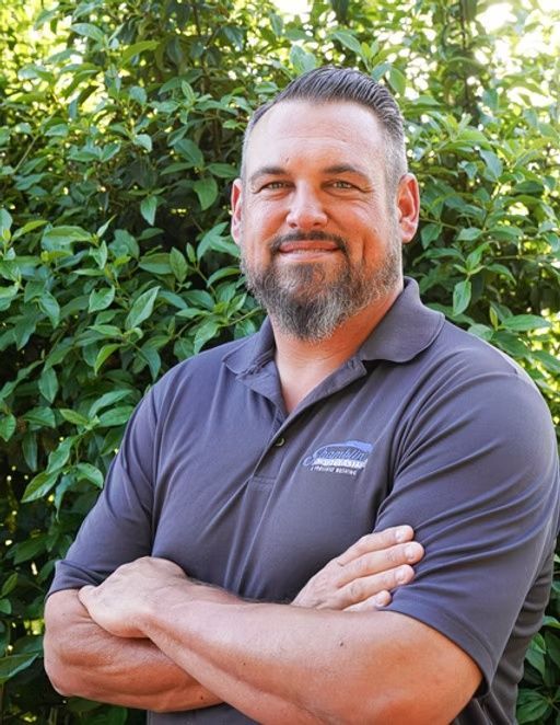 Smiling man with crossed arms in a gray polo, standing in front of green foliage.