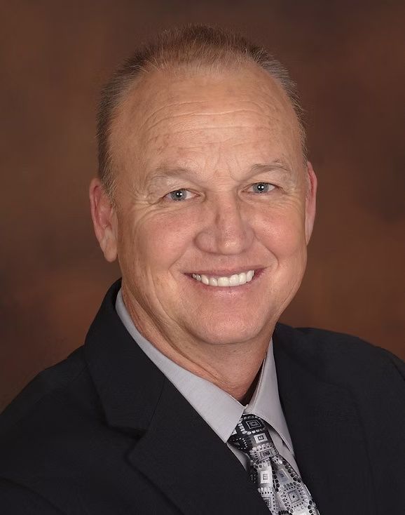 Studio headshot of a smiling man in a dark suit and patterned tie against a brown background