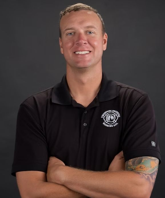 Person in a black polo shirt with a logo, arms crossed, smiling against a dark gray background