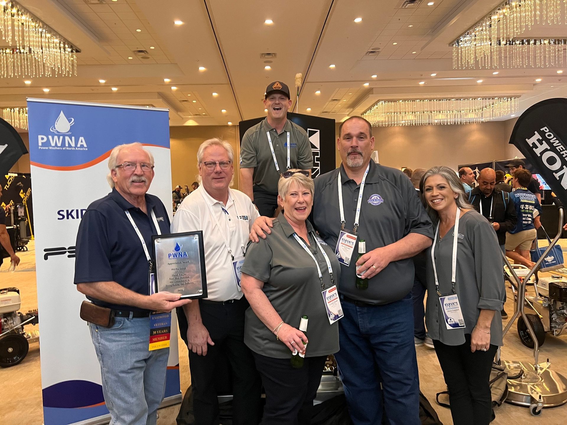 Group of five at a trade show booth with a blue banner and trophy, smiling indoors under bright lights.