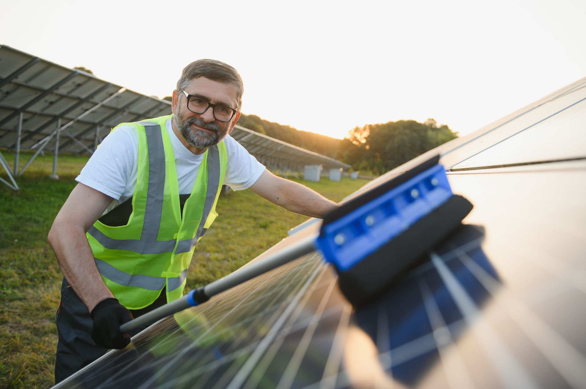 Worker cleaning solar panels with a blue brush outdoors at sunset