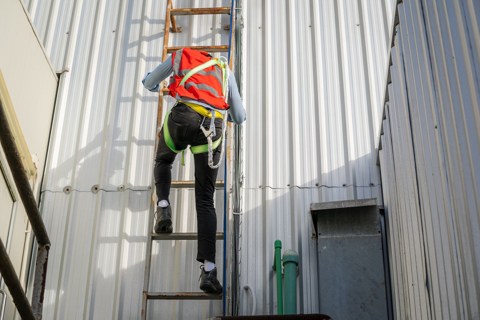 Worker in orange safety vest climbing a ladder against a metal wall on an industrial building