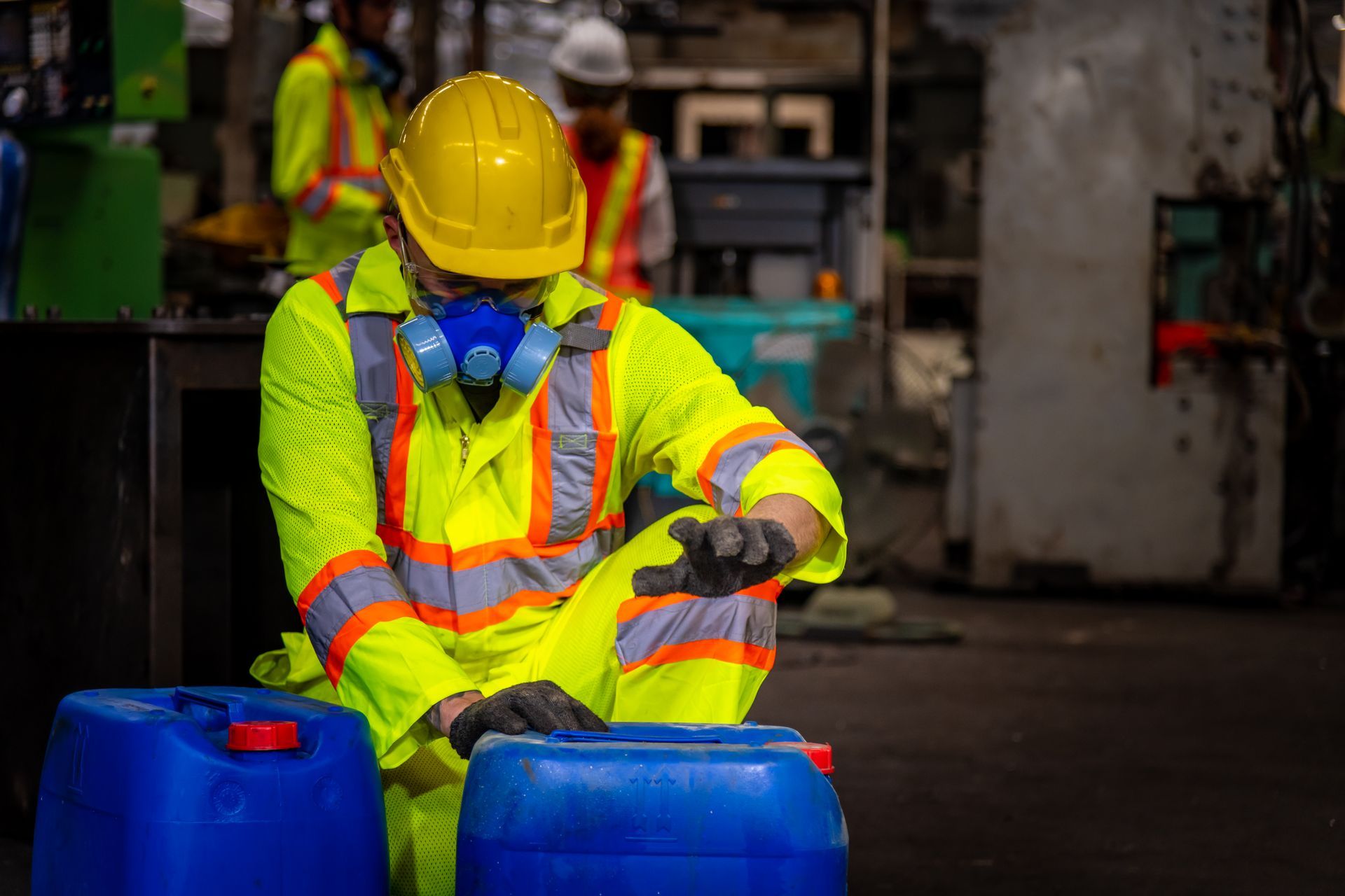 Worker in yellow safety gear and respirator handling blue chemical drums in an industrial setting