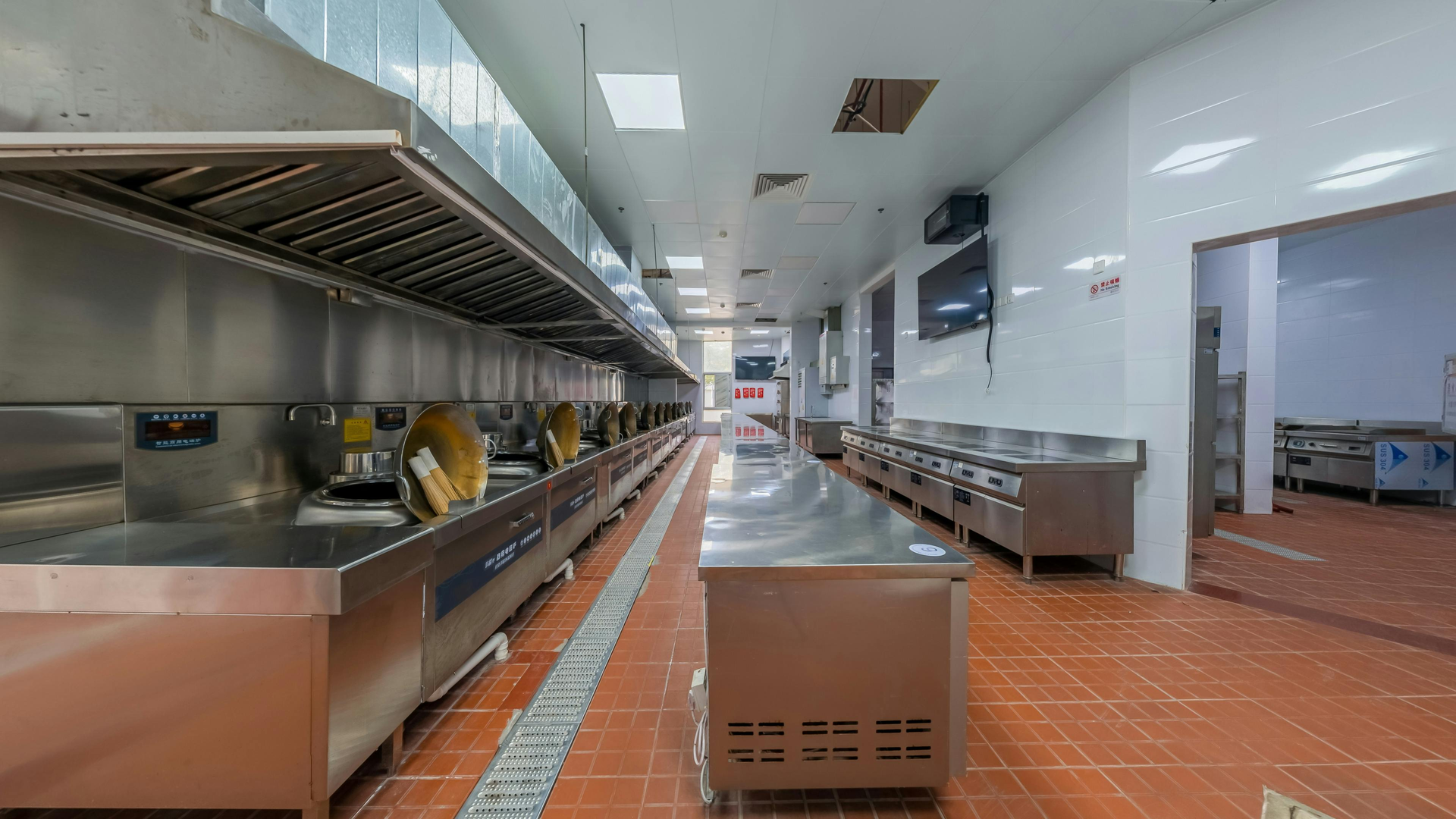 Empty commercial kitchen with stainless steel counters and orange tile floor, viewed down a long aisle