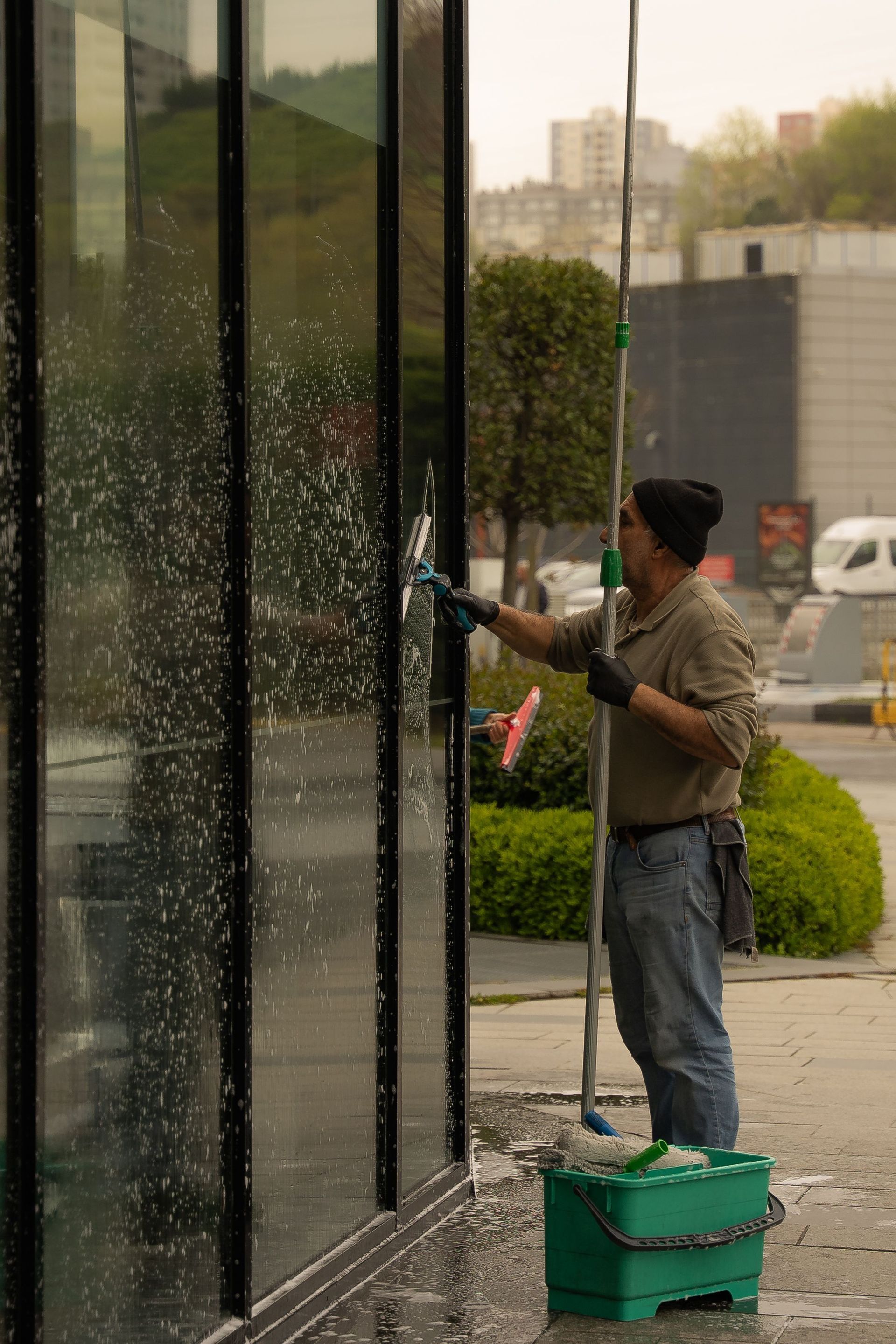 Person cleaning tall glass windows outdoors with a squeegee and green bucket on the sidewalk.
