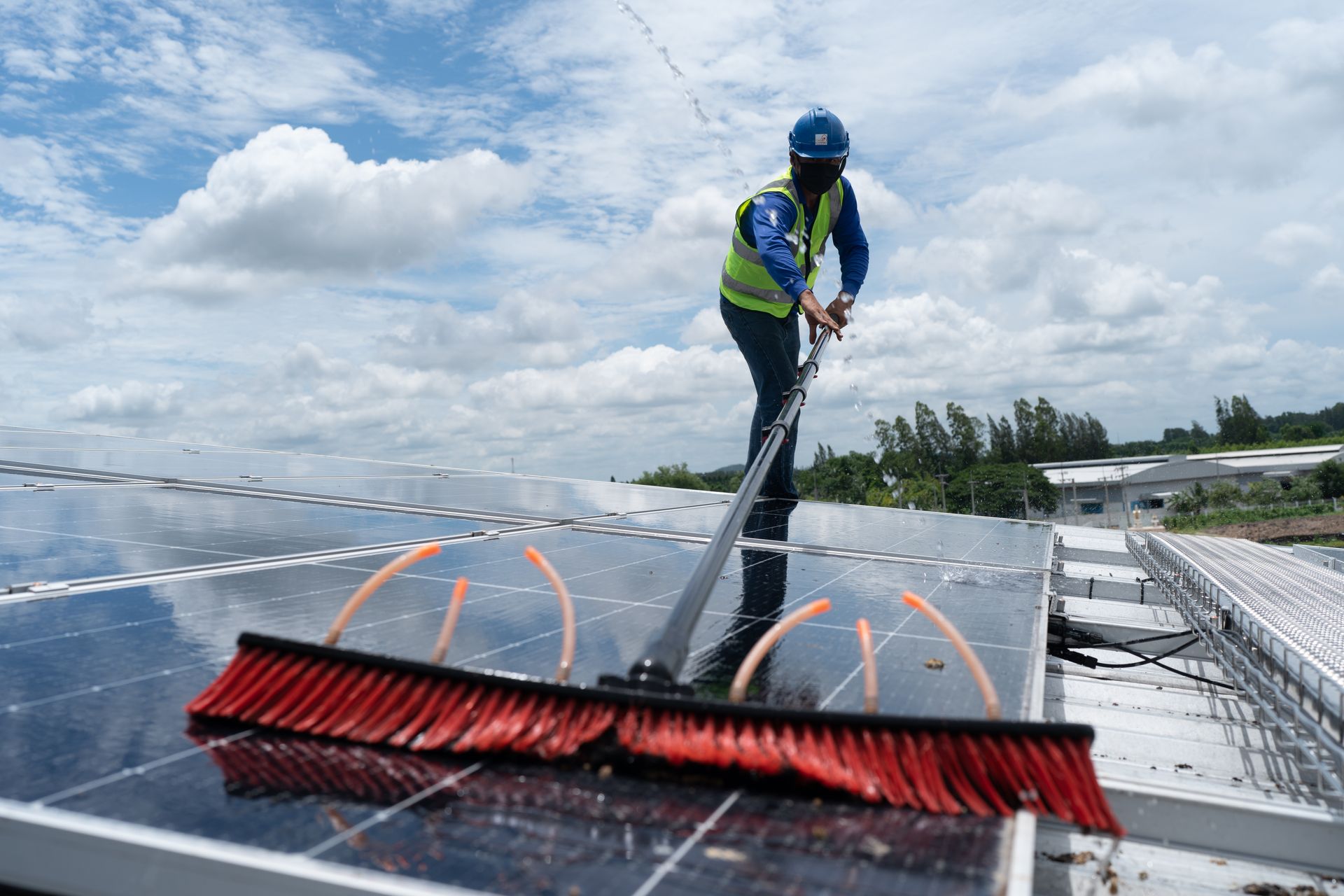 Worker cleaning solar panels with a long brush on a rooftop under a cloudy sky