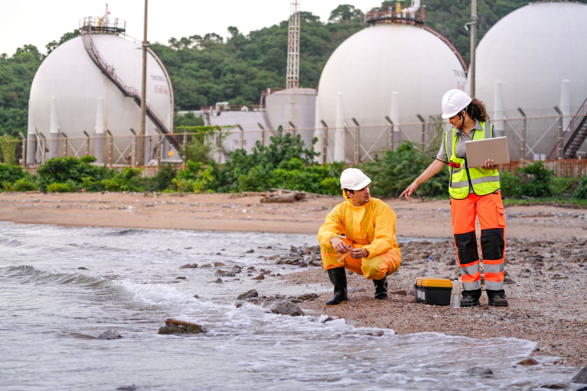 Workers in safety gear sampling water beside an industrial shoreline with storage tanks in the background