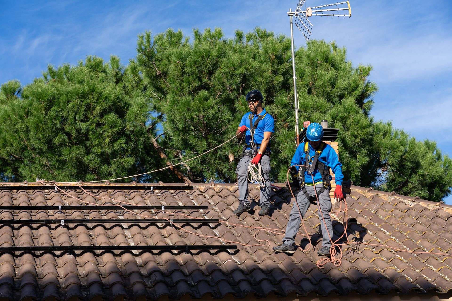 Two roofers in blue gear repair a tiled roof near trees and an antenna.