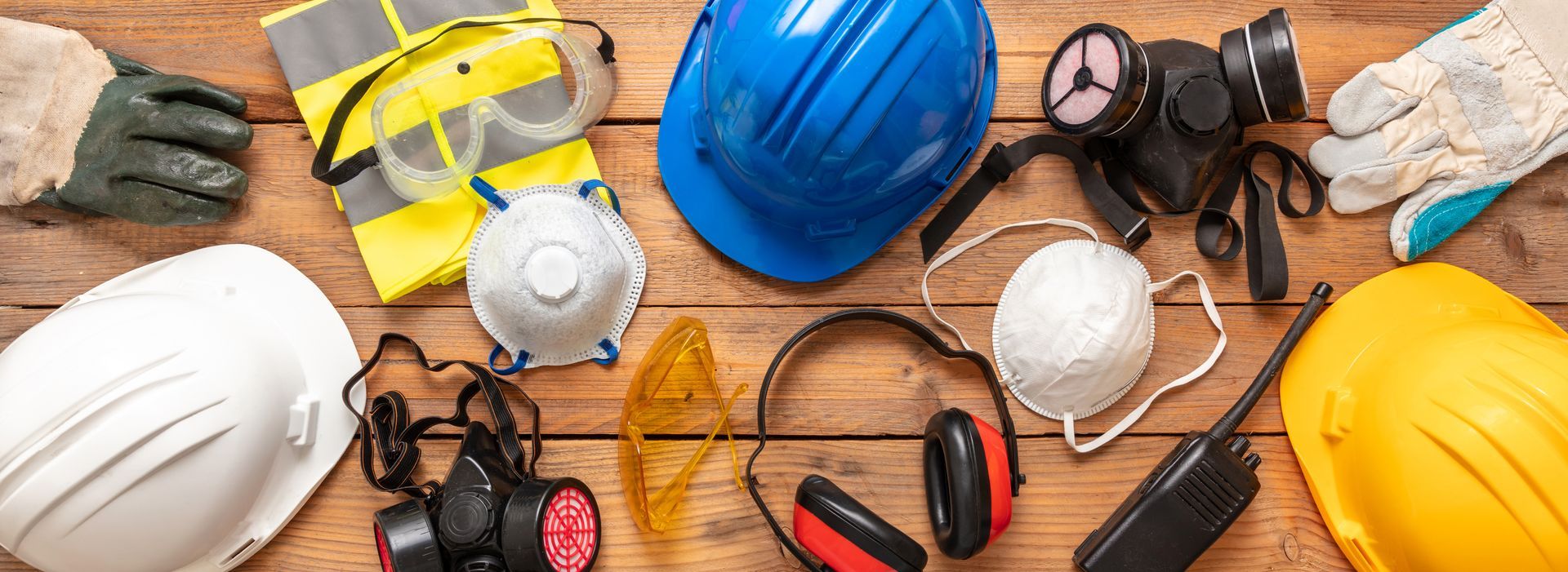 Construction safety gear on wooden surface: hard hats, goggles, respirators, gloves, and ear protection.