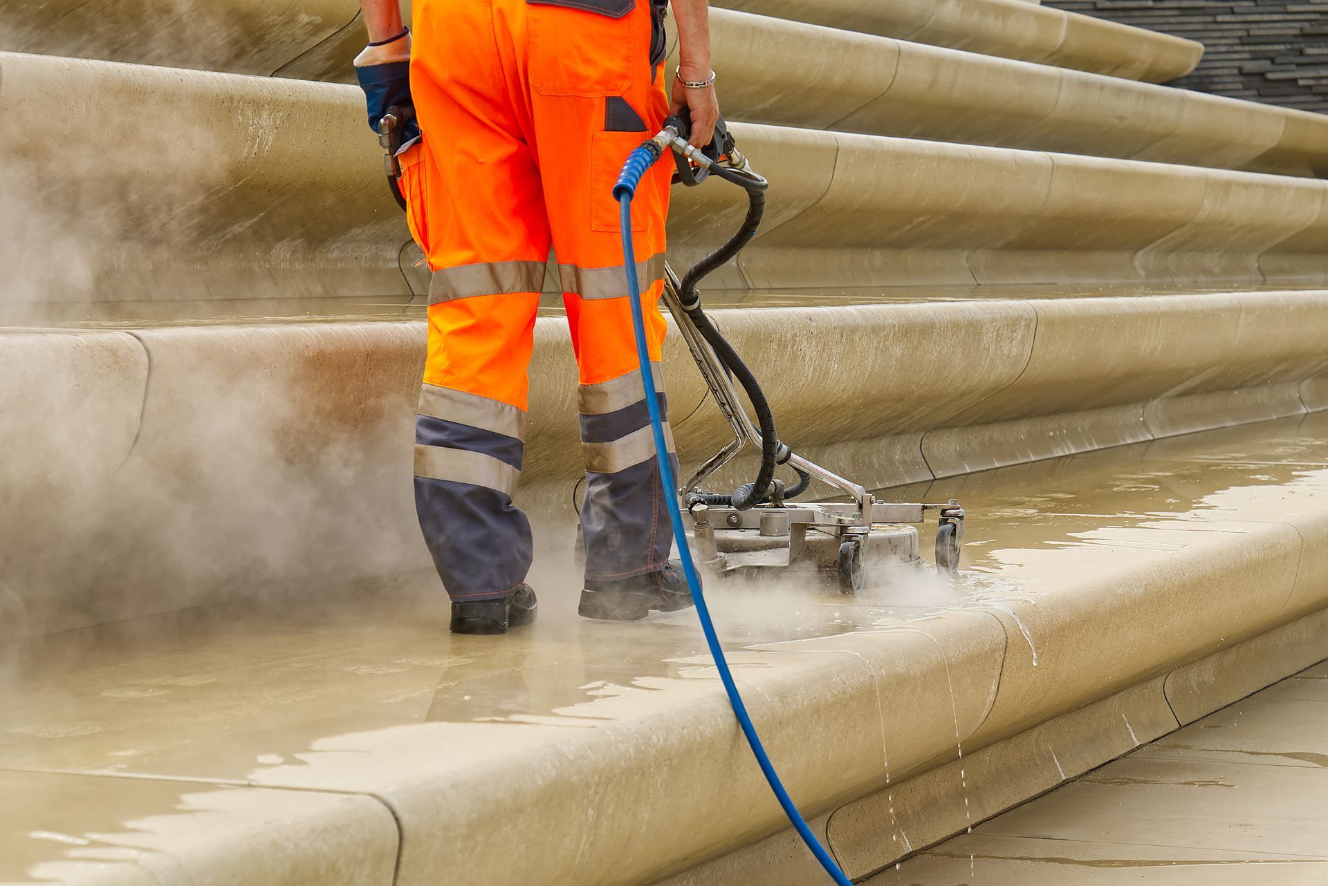 Worker pressure-washing tan stone steps with a blue hose and orange safety gear
