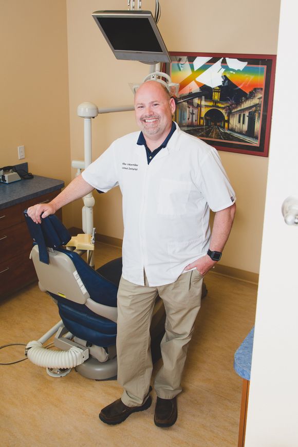Man standing beside a dental chair in a clinic room, smiling, with equipment and artwork on the wall.