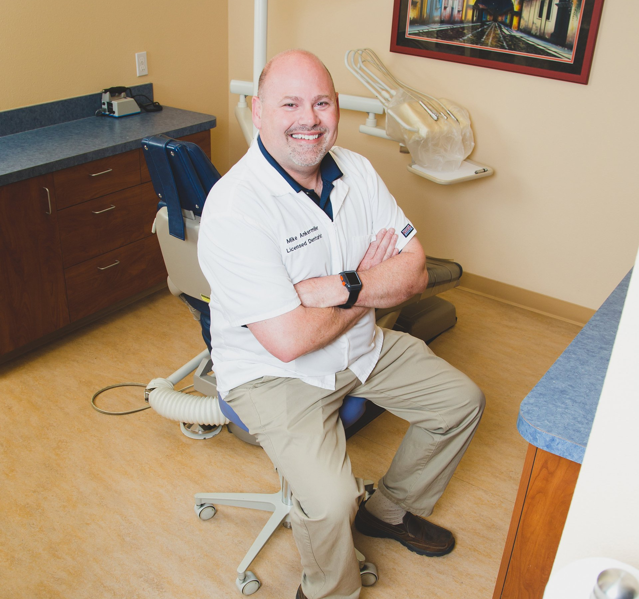 Smiling man seated on a stool in a dental exam room, arms crossed near the dental chair.