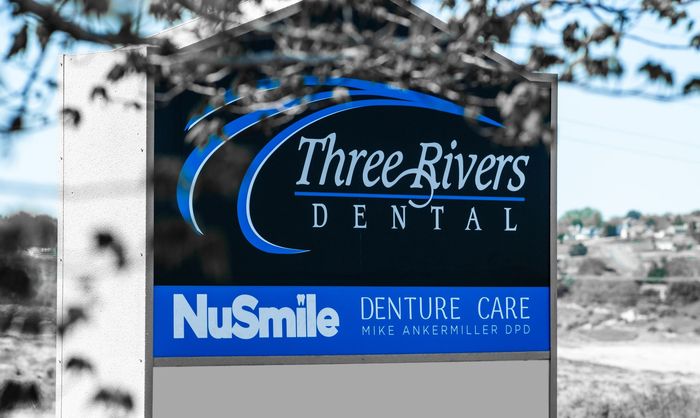 Three Rivers Dental sign with blue NUSmile Denture Care logo in front of a snowy roadside