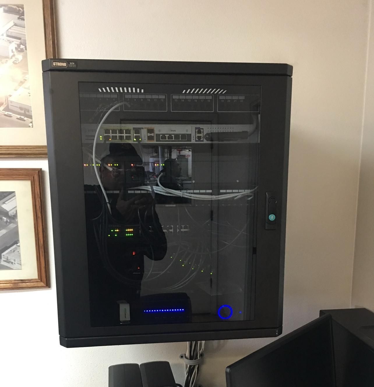 Black wall-mounted server cabinet with glass door and lit electronics inside, in an office corner.