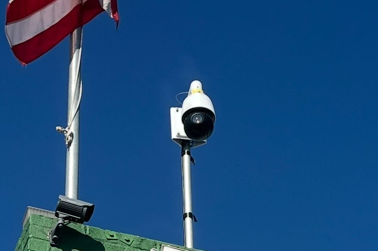 A white security camera on a pole beside an American flag against a blue sky