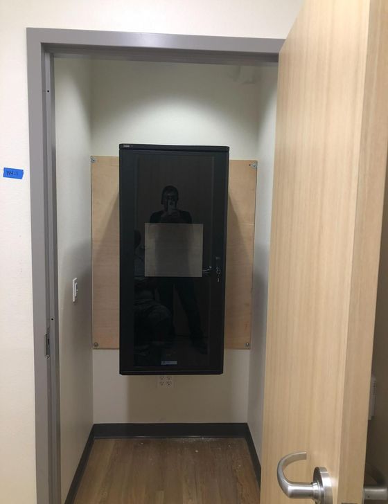 Narrow restroom with a black wall-mounted dispenser or mirror between wood-paneled walls, door open.