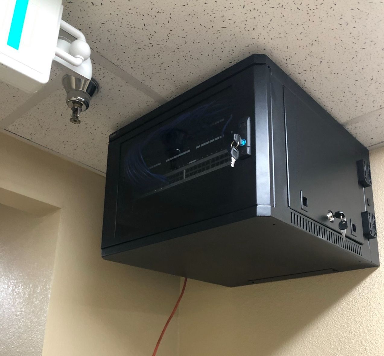 Black wall-mounted AV/IT cabinet near the ceiling, with cables and a small device attached underneath.