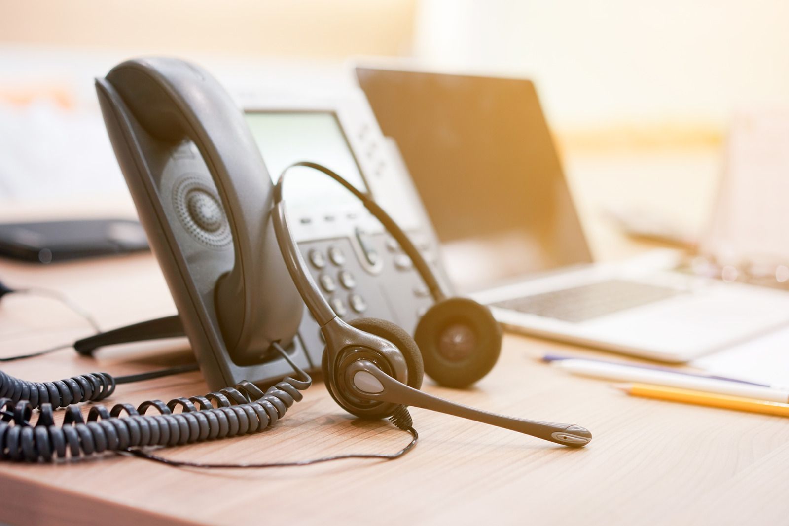 Office desk with corded phone headset, laptop, and pencil in warm light
