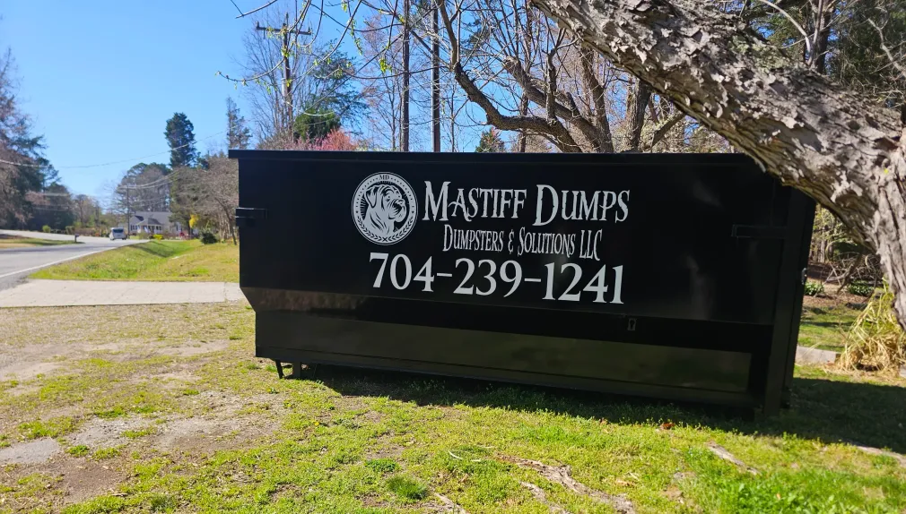 A black dumpster is sitting in the grass next to a tree.