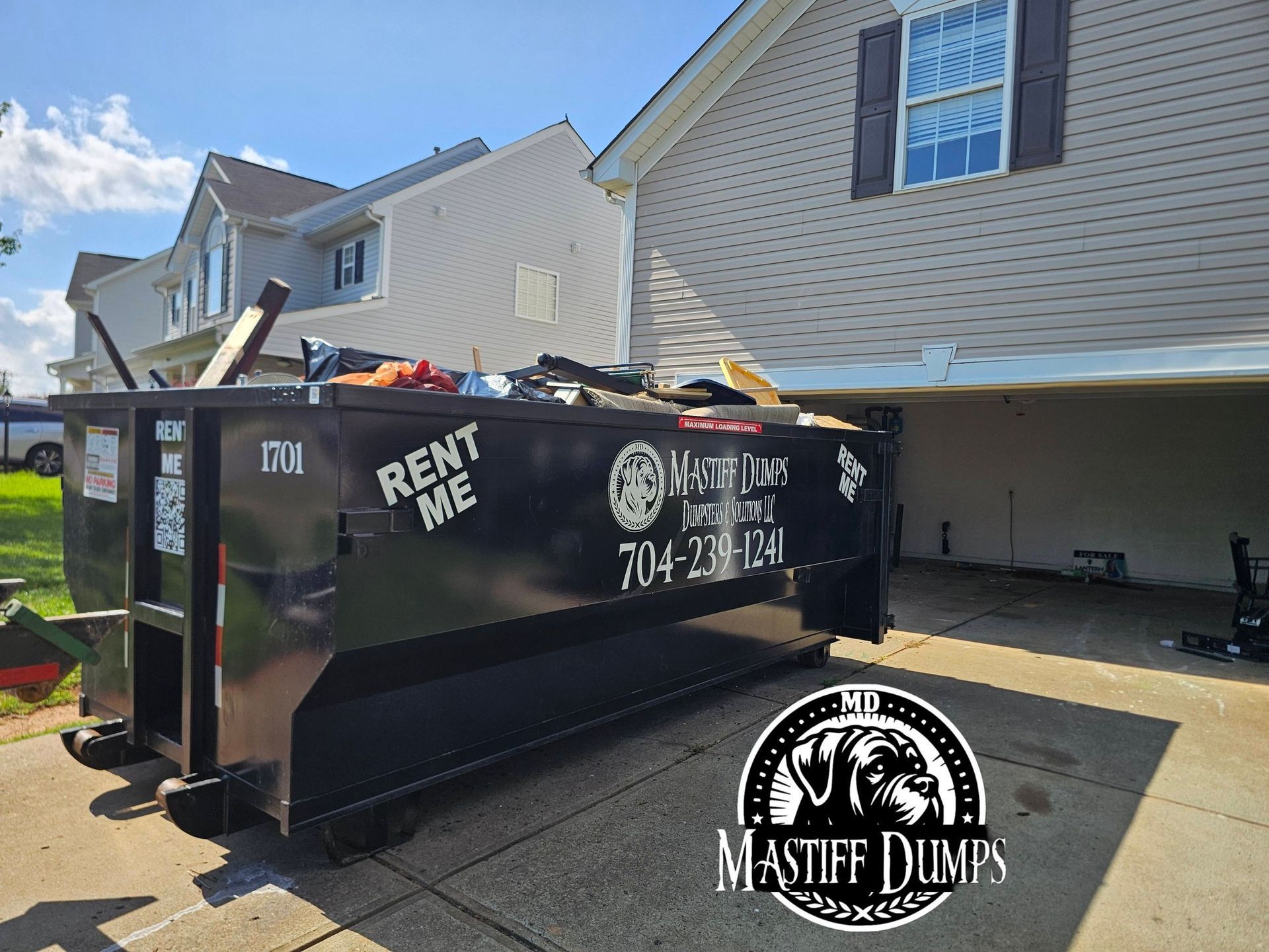 A black dumpster is sitting in the grass next to a tree.