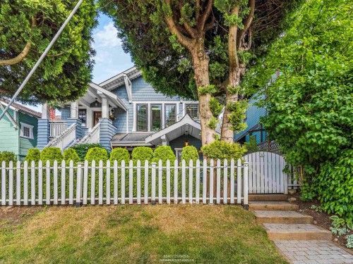 White picket-fenced house with blue siding, trees, and a front lawn on a sunny day