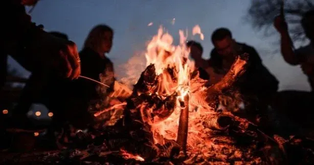 People gathered around a glowing campfire at dusk, with flames and embers lighting the scene.