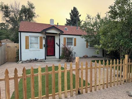 Small white house with red door, brown roof, and a picket fence at sunset