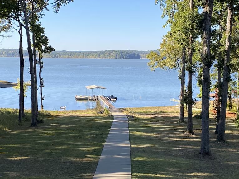 Lakeside dock with a concrete path through trees to blue water and a distant shoreline.