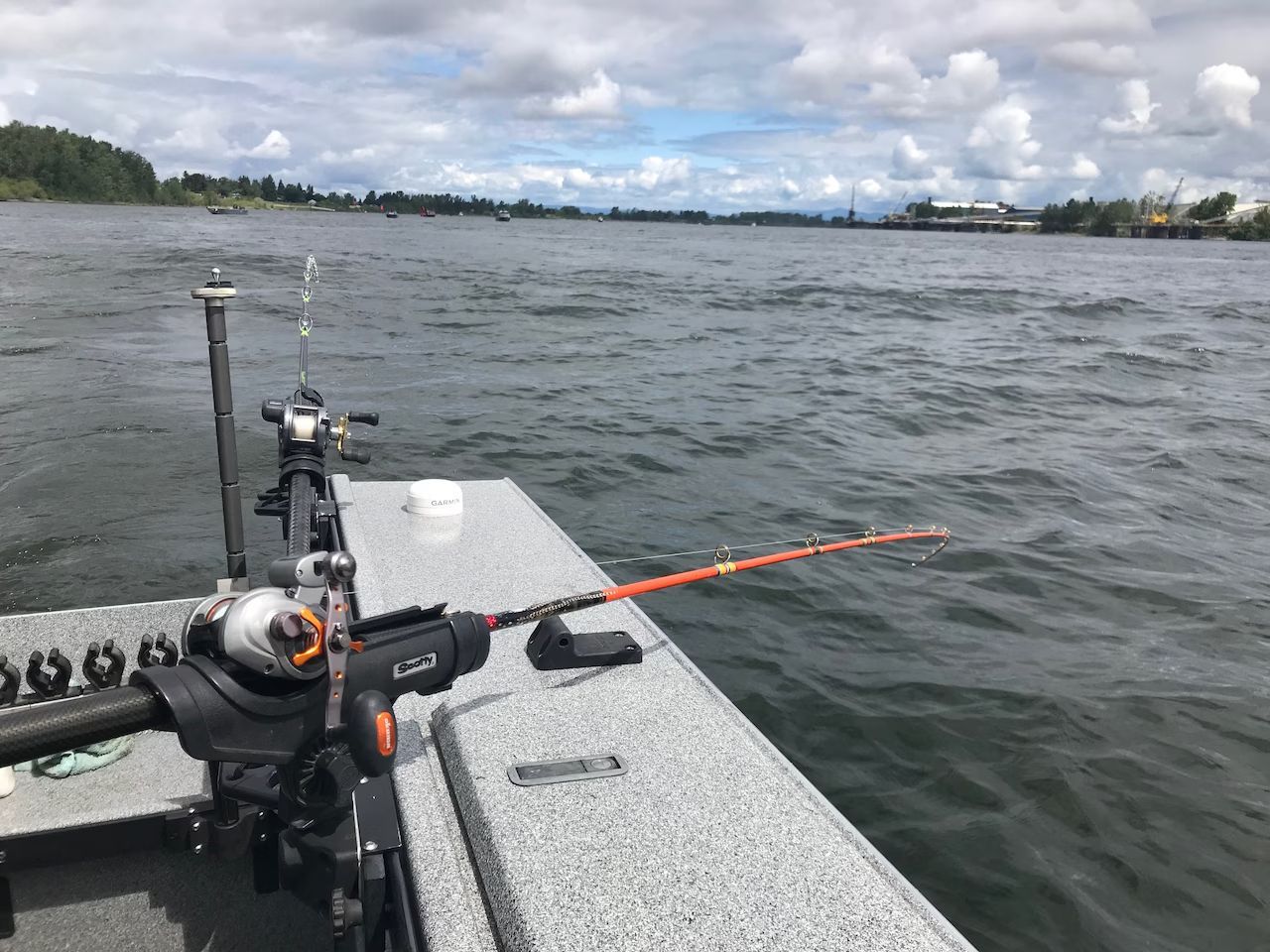 Fishing rod on a boat over choppy water near a wooded shoreline under cloudy skies