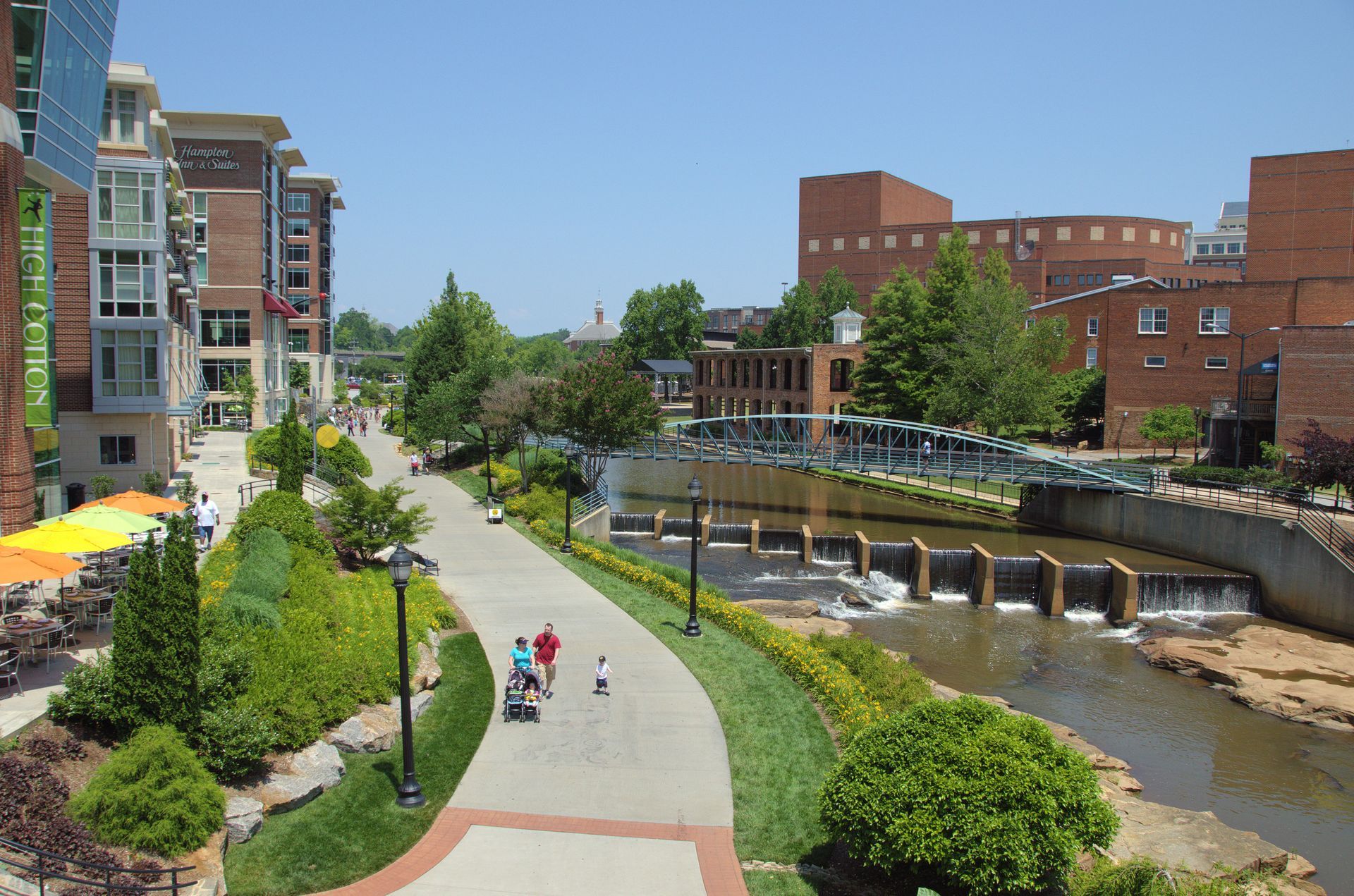Sunny riverside walkway beside modern buildings, with people strolling and a low water cascade.