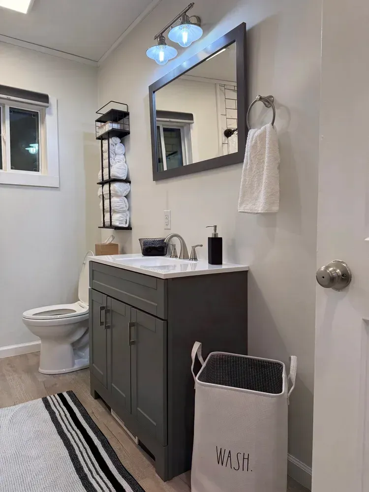 Small bathroom with gray vanity, white sink, mirror, toilet, striped rug, and a towel rack.