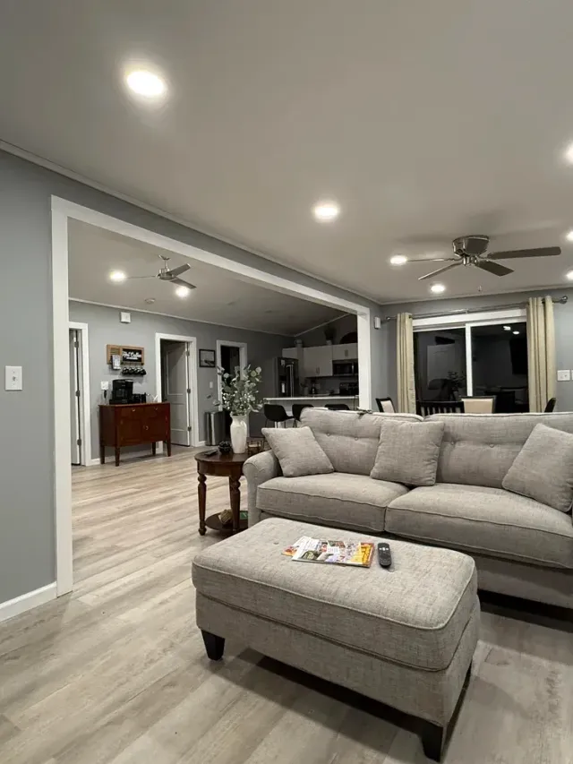 Modern open-concept living room with gray sectional, ottoman, recessed lights, and adjacent dining area