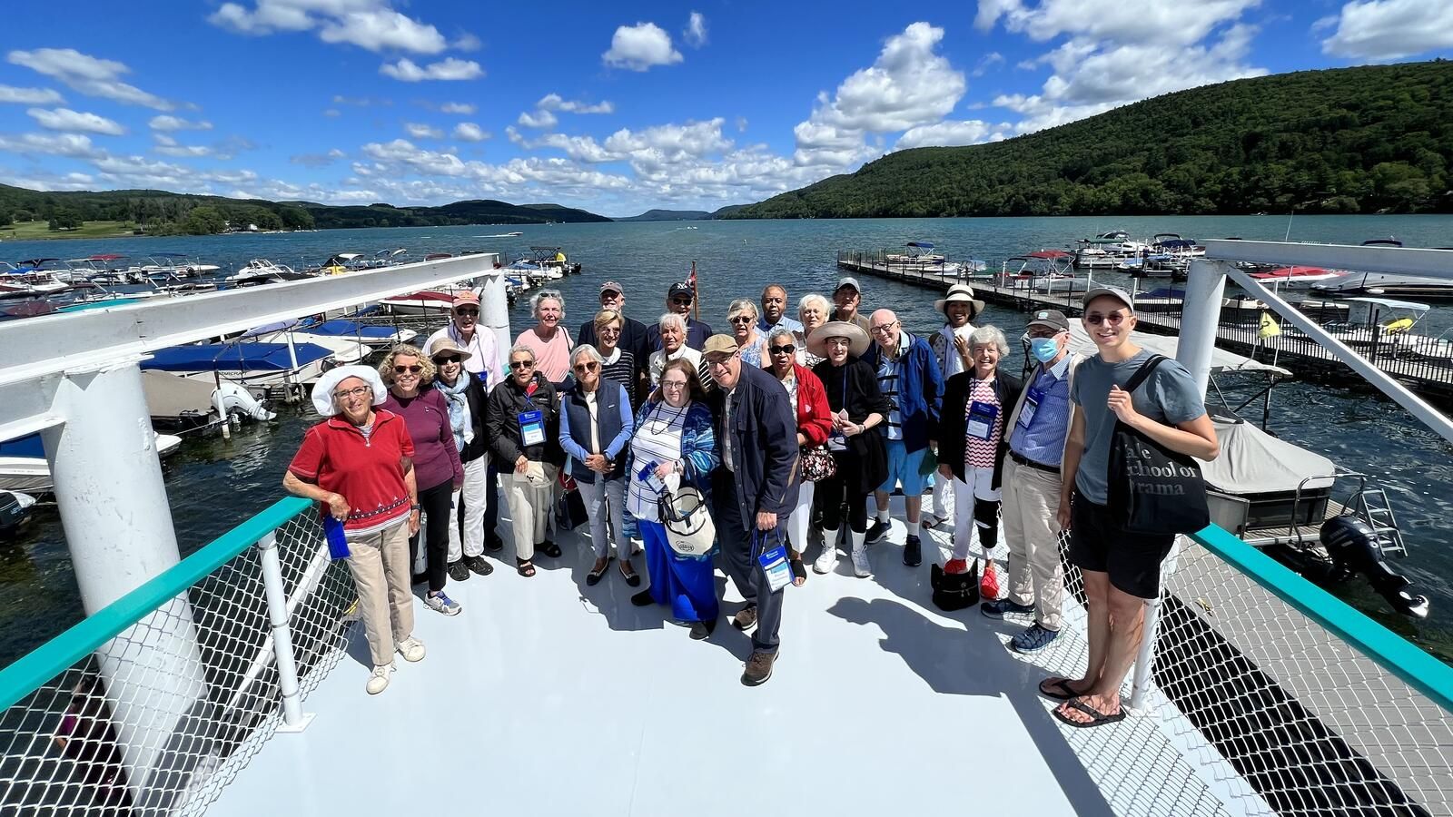 Group of people standing on a docked boat deck with water, mountains, and a blue sky in the background